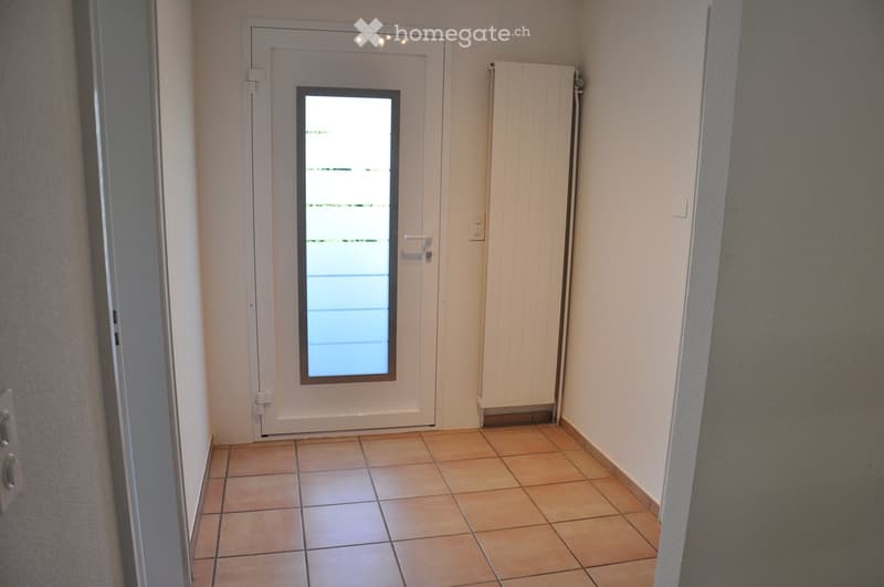 A narrow hallway with tiled flooring, a white door with a glass panel, and a radiator on the wall.