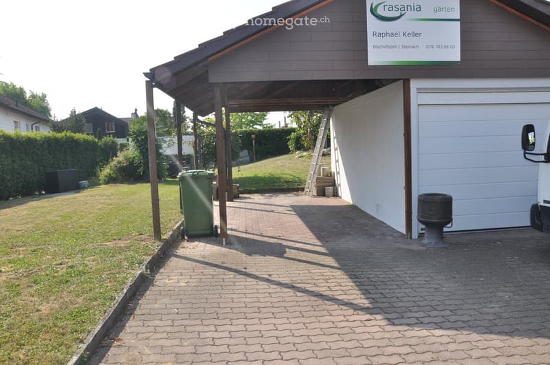 A paved driveway leads to a covered garage area with a sign above. A green trash bin is visible on the left, and a barbecue grill is near the garage. The surrounding area includes a well-maintained lawn and hedges.