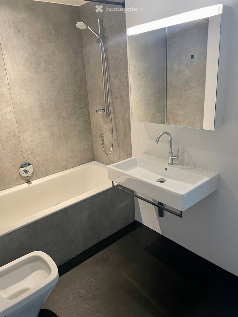 Modern bathroom with a bathtub, wall-mounted sink, mirror, and showerhead. The walls are finished in a textured grey material, and the floor is dark.