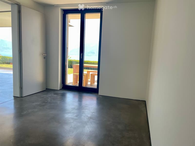 Empty room with concrete floor, white walls, large sliding glass door leading to outdoor area with wooden furniture visible outside.