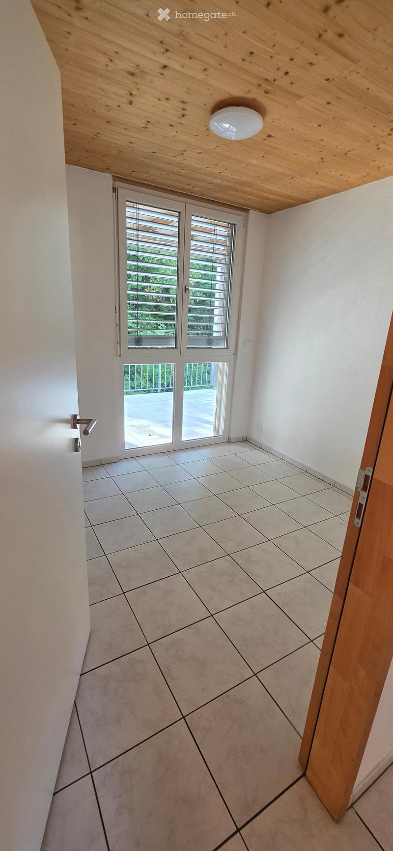 Empty hallway with tiled floor, white walls, wooden ceiling, and sliding glass doors leading to a balcony or outdoor area.