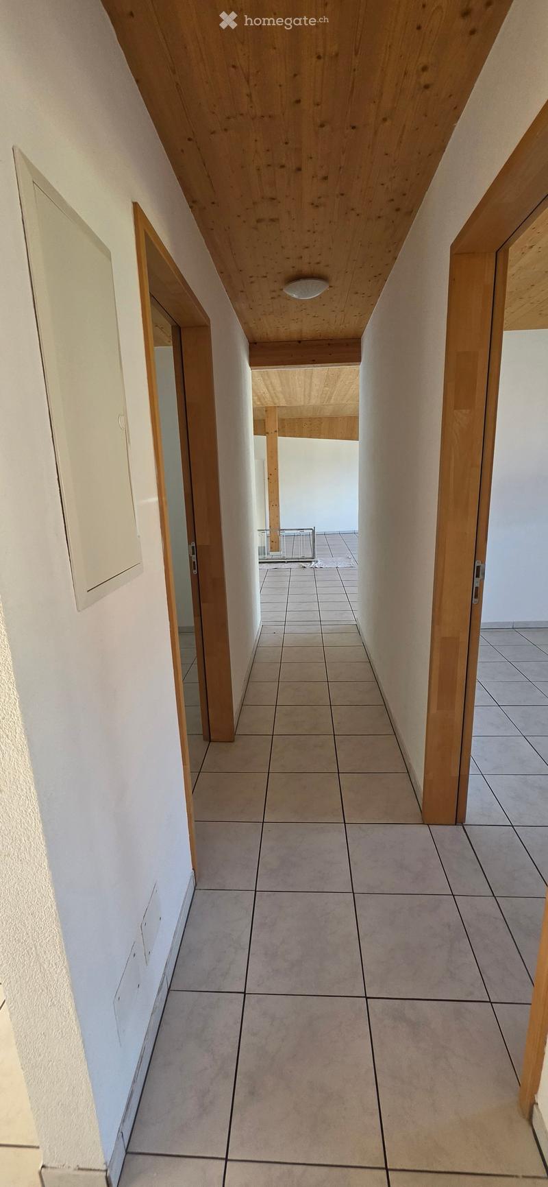 A modern hallway with tiled flooring, white walls, and wooden structural elements. The ceiling features exposed wooden beams and recessed lighting. Doors are visible on both sides, leading to other rooms.