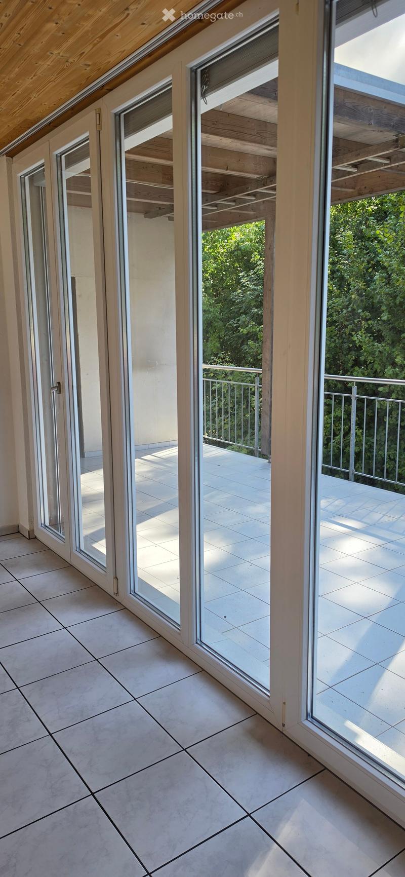 View of a modern balcony area with large glass doors, wooden ceiling beams, and a tiled floor, leading to an outdoor space with a metal railing and greenery visible beyond.