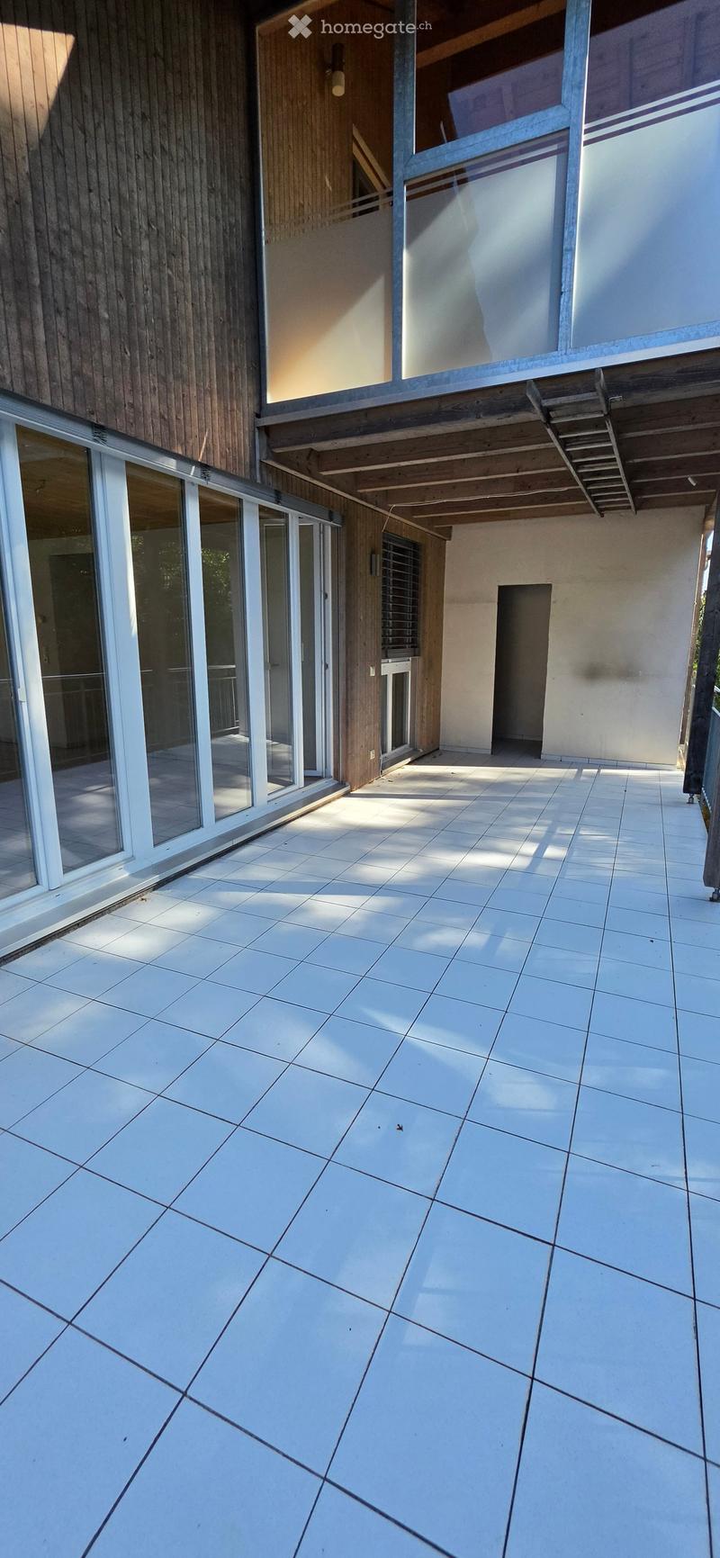 Modern outdoor patio with large white tiles, wooden decking, and large glass sliding doors leading to the interior.