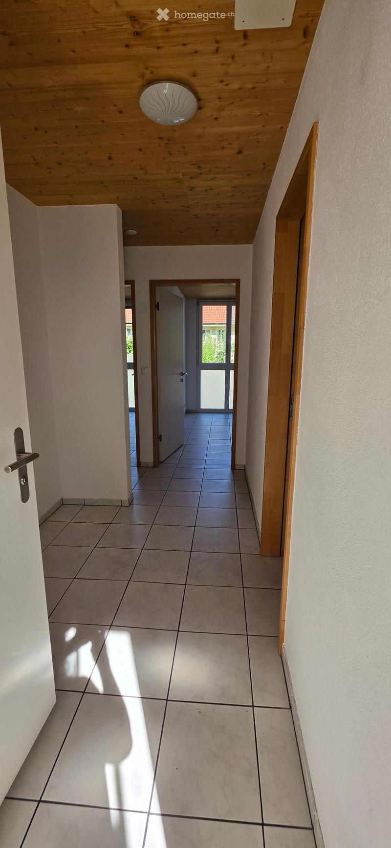 Empty hallway with tiled floor, wooden ceiling, and white walls. Features a door on the left and an open doorway leading to another room on the right.