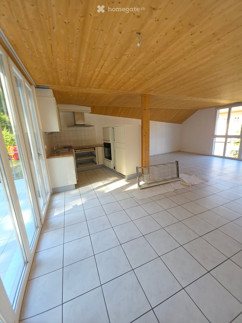 Empty room with white tiled floor, white cabinets, and a wooden slanted ceiling. The room has large windows and a small kitchen area with a sink and stove.