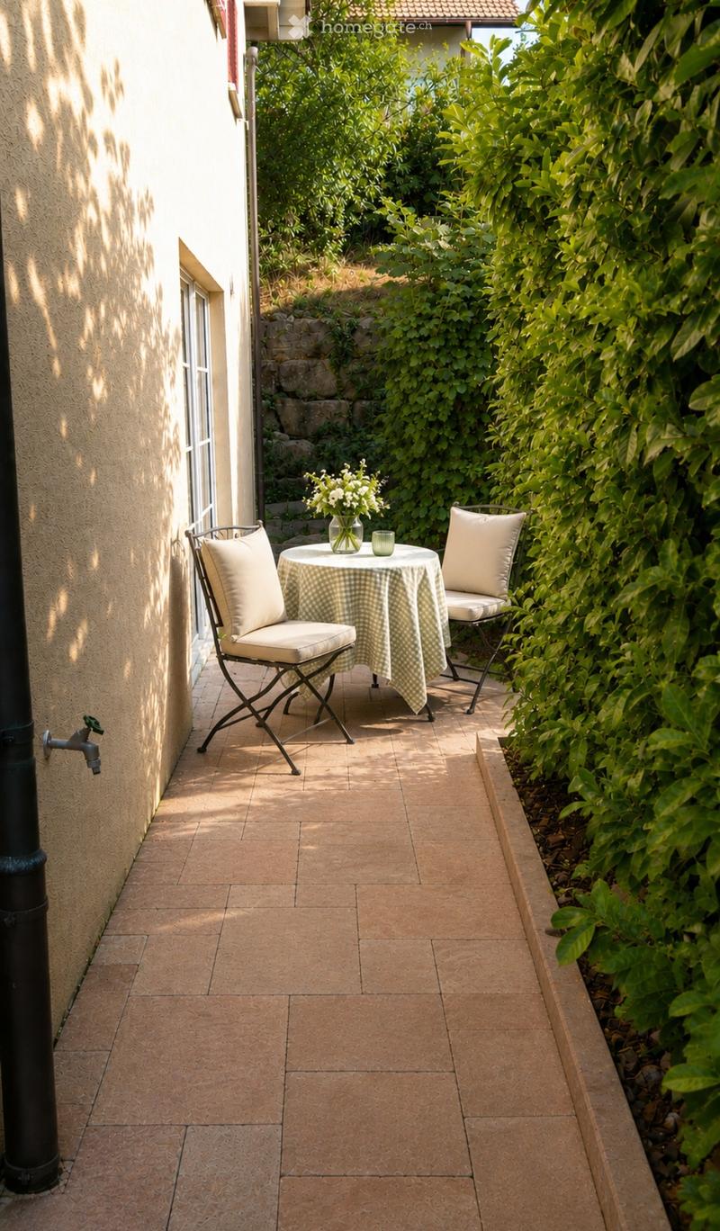 Balcony with tiled floor, small round table with checkered tablecloth, two matching chairs, vase with flowers, surrounded by lush greenery.