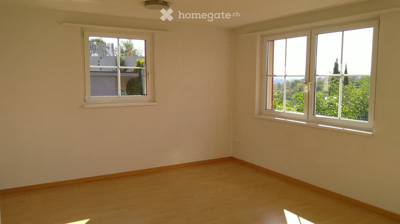 Empty room with light-colored walls, wooden flooring, and two windows offering views of greenery and neighboring buildings.