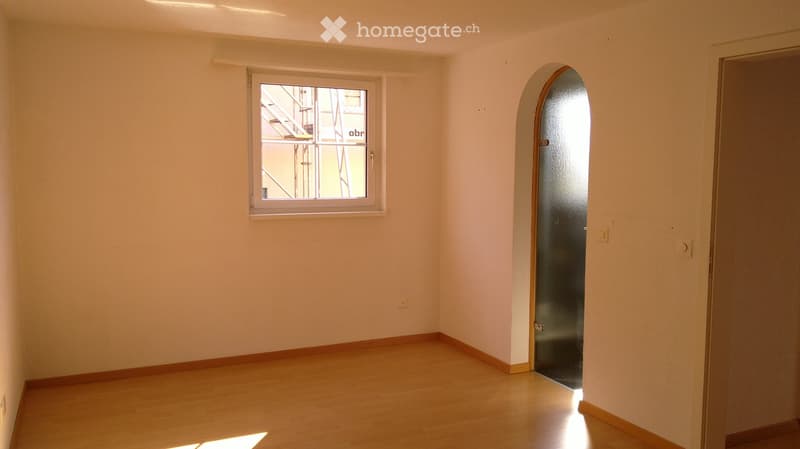 Empty room with light-colored walls, wooden flooring, and a single window with a view of a staircase outside. The room has a door with a frosted glass panel on one side.