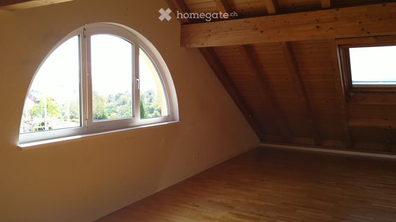 Empty attic room with wooden slanted ceilings, light wooden flooring, and large arched window offering a view of greenery outside.