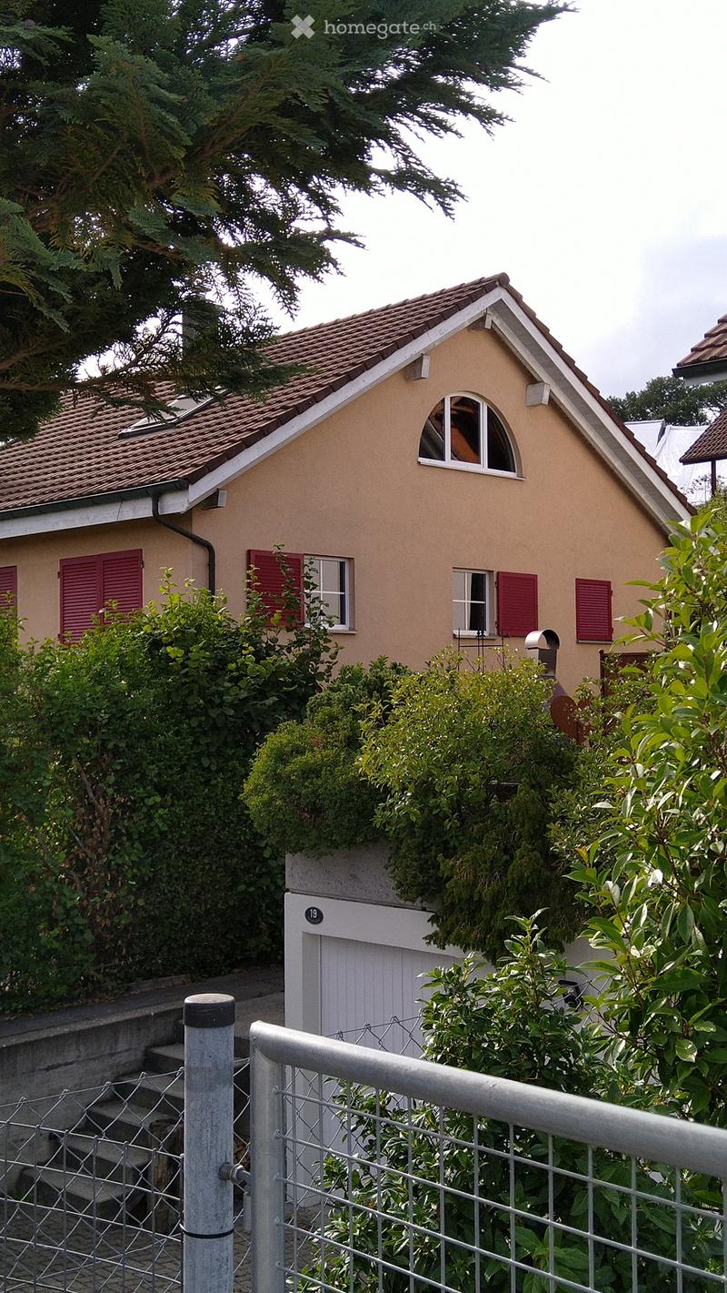 A small, single-story house with a tiled roof, beige exterior, red shutters, and a white garage door. The house is surrounded by greenery and has a metal fence in the foreground.