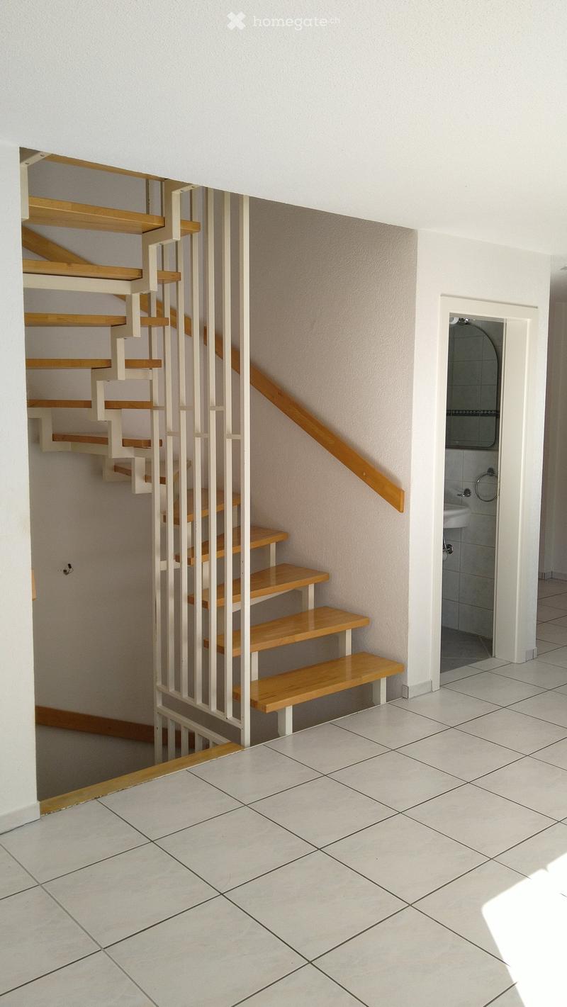 A modern staircase with wooden steps and white railings leads up, adjacent to a small tiled bathroom with a sink and mirror.