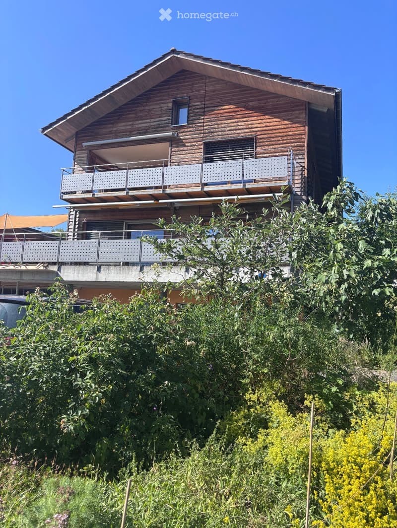 Modern two-story house with wooden exterior, multiple balconies, and lush greenery in the foreground.
