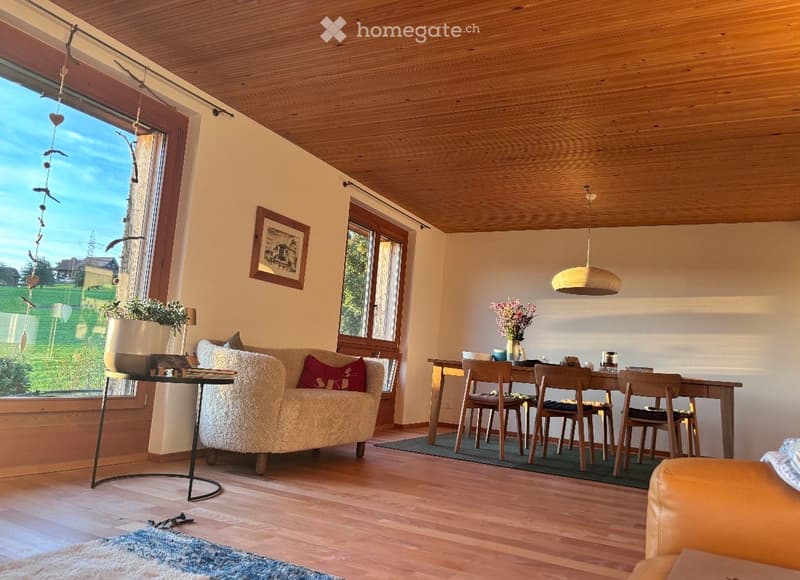 Bright living and dining area with wooden floors, large windows offering a view, a cream-colored sofa, a coffee table, a dining table with chairs, and decorative items including a vase with flowers and a picture frame.