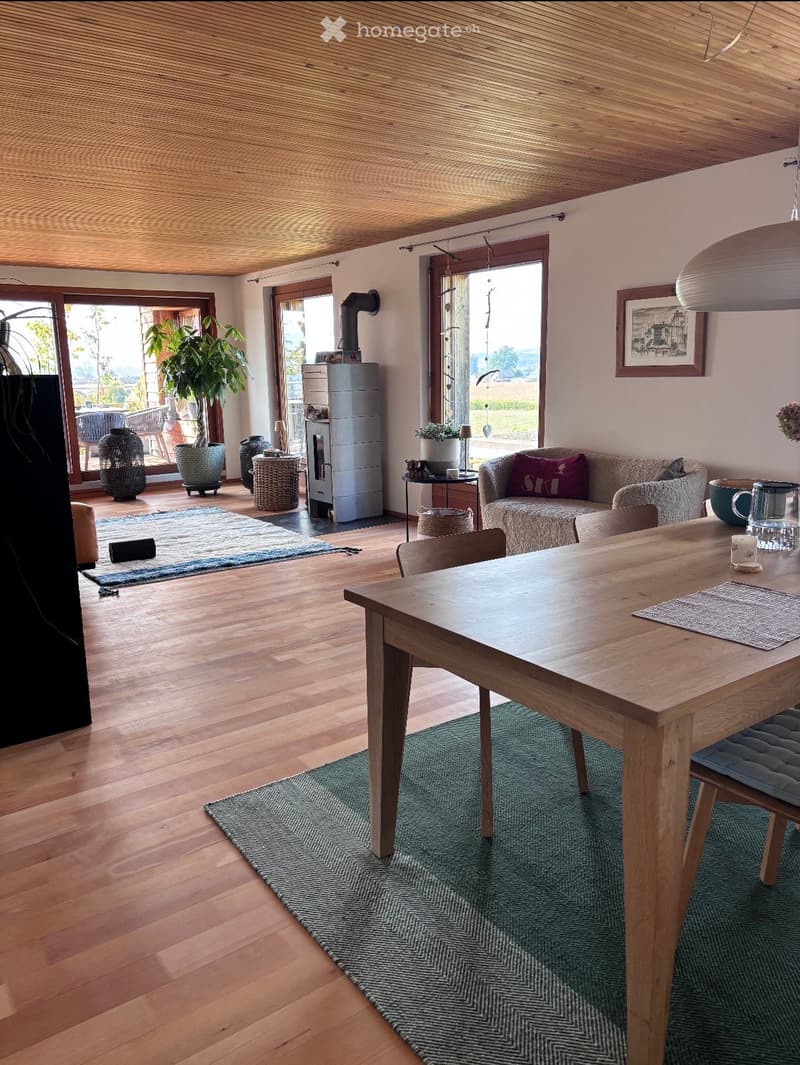 Bright, open living and dining area with wooden floors and ceiling, large windows, a sofa, dining table, and a wood-burning stove.