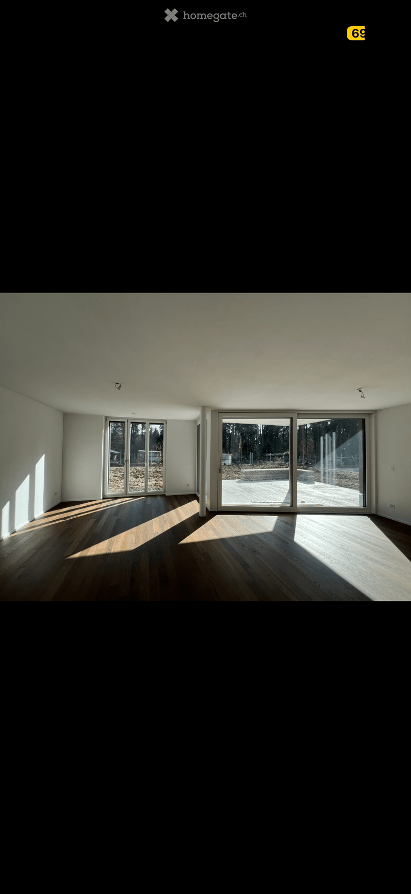 Bright, empty room with wooden flooring and large sliding glass doors leading to an outdoor deck. Sunlight streams in, casting shadows on the floor.