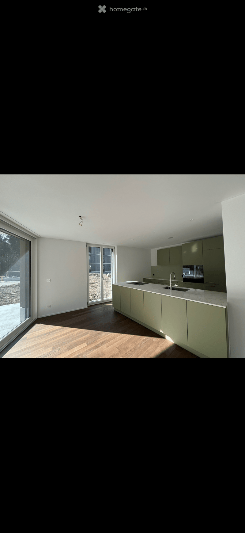 Modern kitchen with light green cabinets, marble countertop, and wooden flooring. Features a large window and sliding glass door leading to an outdoor area.