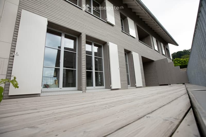 Modern two-story house with grey siding, large windows, and white shutters. The deck in the foreground is made of light-colored wood.