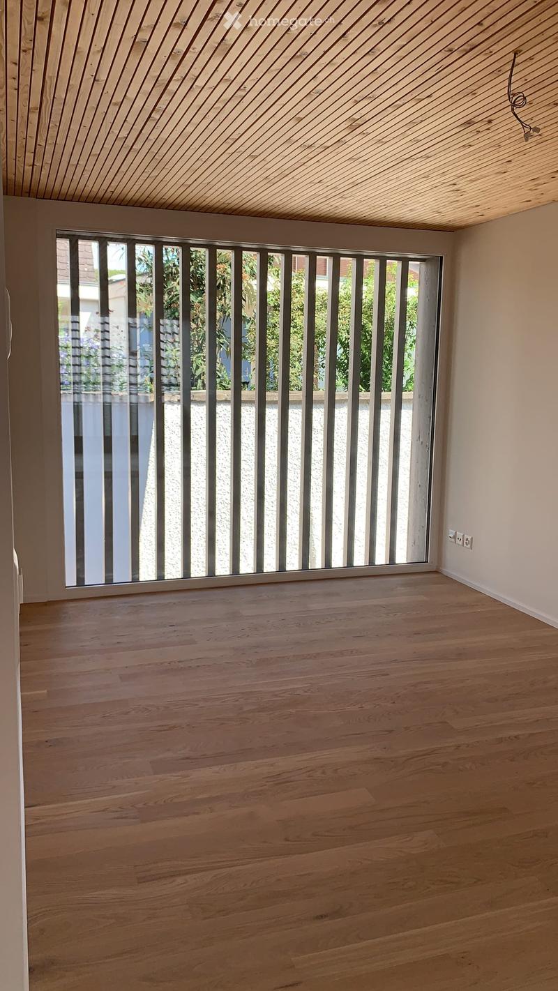 Empty room with wooden flooring, large window with metal bars, and white walls.