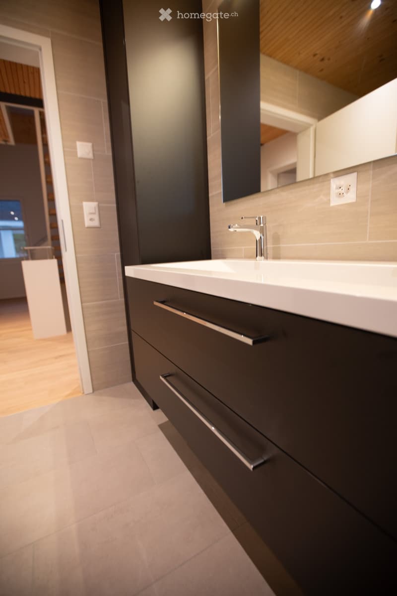 Modern bathroom with a white sink, dark cabinet, and sleek faucet. Light-colored tiles and wooden flooring.