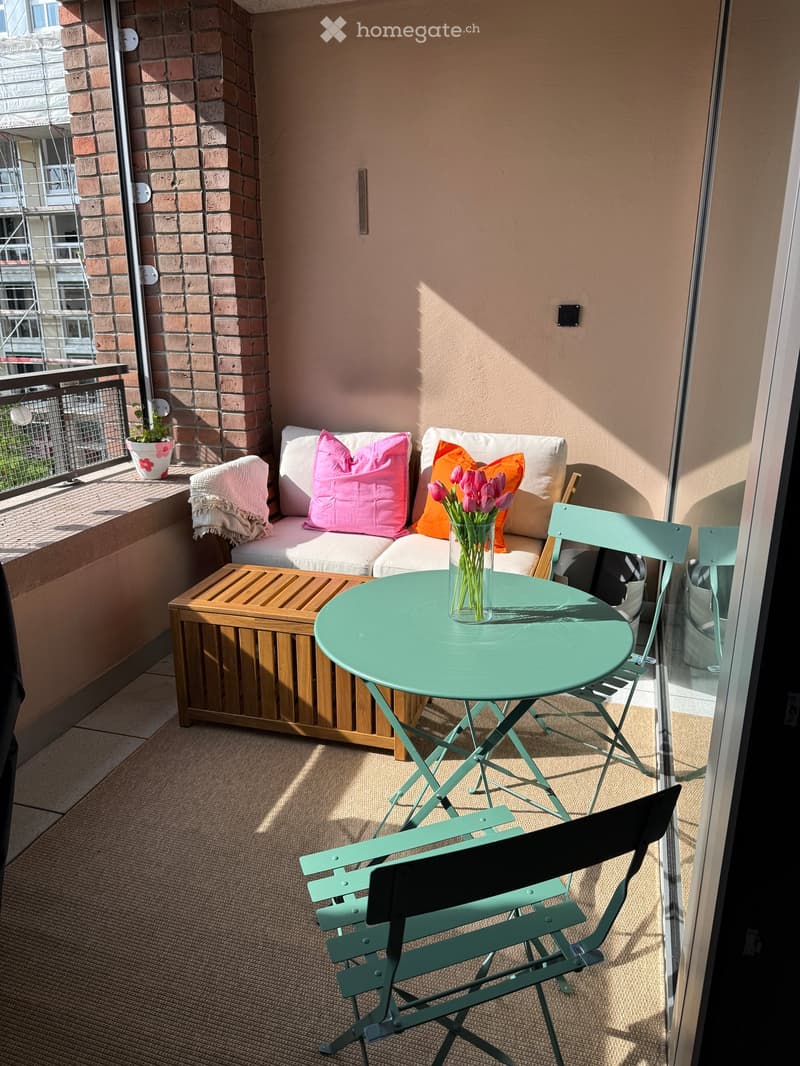 A small balcony with a white sofa, colorful cushions, a round green table, two green folding chairs, a vase with tulips, and a wooden storage box.