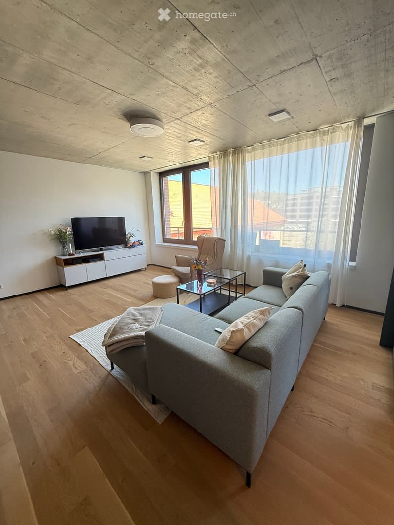 Modern living room with light wooden flooring, grey sectional sofa, coffee table, armchair, ottoman, TV stand, and decorative vases.