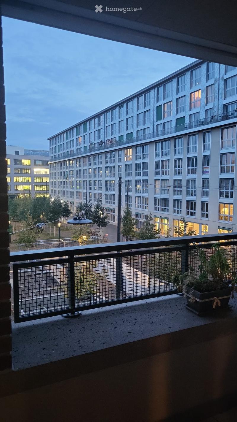 Balcony with metal railing, potted plant, overlooking a modern apartment building complex with multiple floors and illuminated windows.