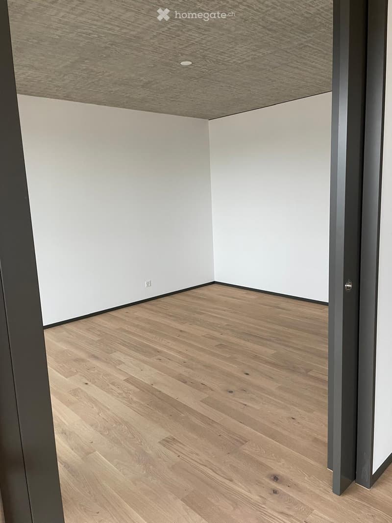 Empty room with light wooden flooring and white walls, featuring a visible ceiling and a door frame.