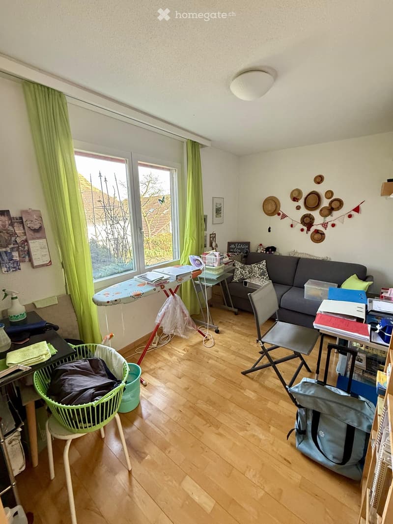 room with wooden floor, large window, couch, table, ironing board, chair, desk, storage boxes, bags, books, and other items