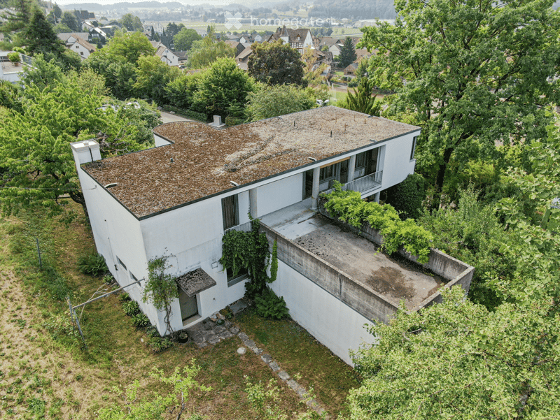 A modern, white, single-story house with a flat roof and large windows, surrounded by lush greenery and trees. The house features a raised ground floor and is situated in a scenic, elevated location with a panoramic view of the surrounding landscape.