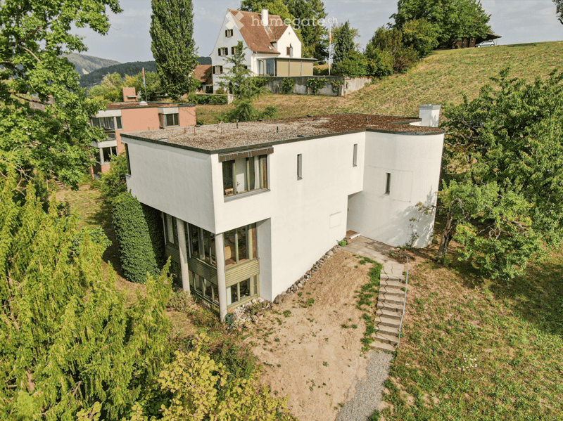 A modern white house with a flat roof and large windows, situated on a hillside surrounded by greenery and other houses.