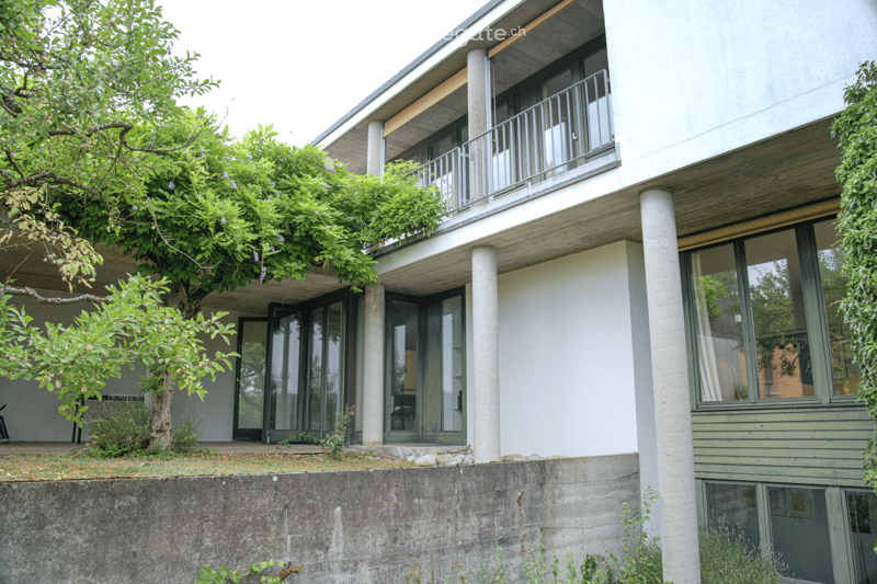 Modern two-story building with white walls, large windows, and a balcony on the upper level. The ground floor has a raised entrance with a small garden area and trees.