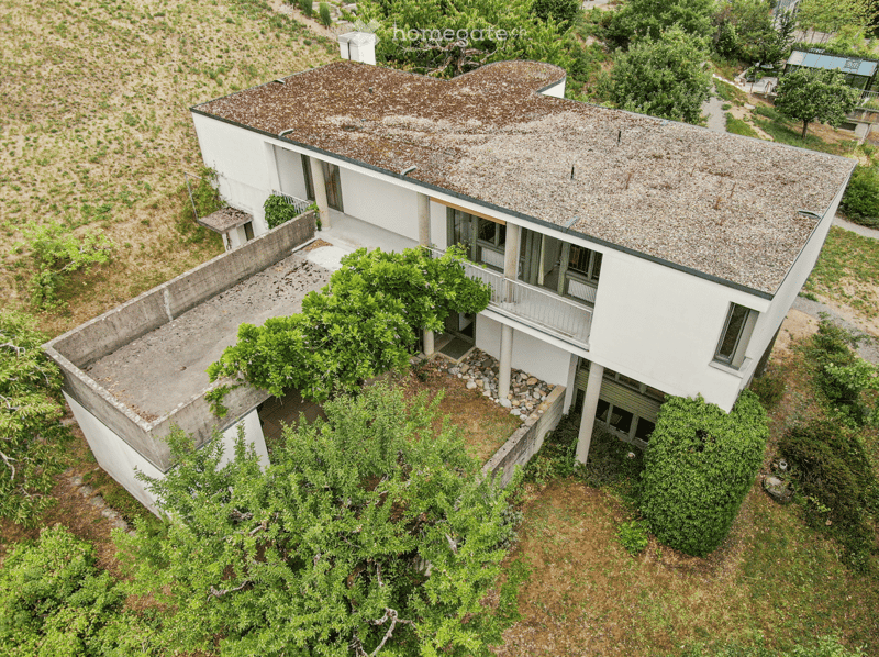 Aerial view of a modern white house with a unique, curved roof and multiple levels, surrounded by greenery and trees.