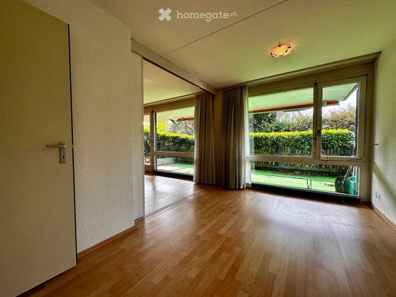 Empty room with wooden flooring, large windows, and sliding glass doors leading to a garden area.