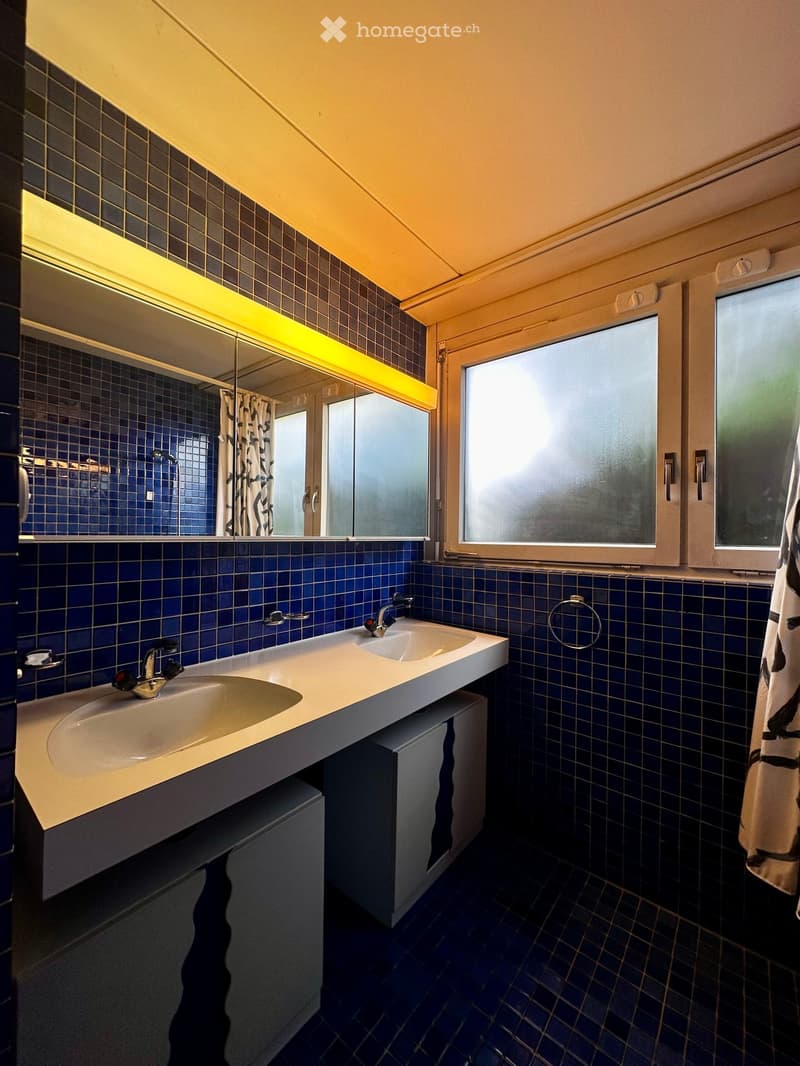 Modern bathroom with blue tiles, double sink, large mirror with neon lighting, and frosted windows.