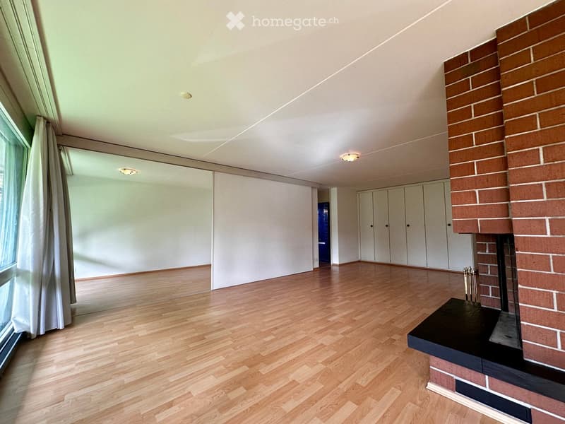 Empty room with wooden flooring, white walls, large window with white curtains, and a brick fireplace with a black mantel.