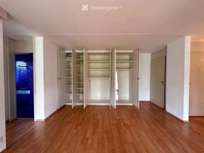 Empty room with wooden flooring, white walls, and built-in open shelving units. A blue-tiled bathroom is visible through an open doorway.