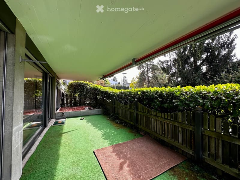 Balcony with green artificial turf, wooden fence, small garden area with rocks and plants, and a red doormat near the entrance.