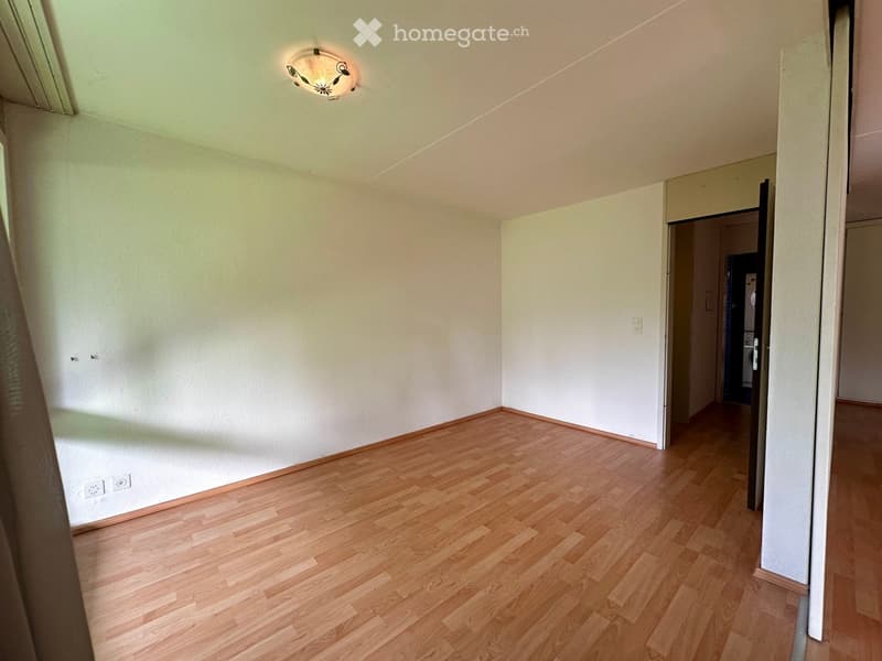 Empty room with light wooden flooring, white walls, and a ceiling light fixture. A doorway leads to another room with a visible washing machine.