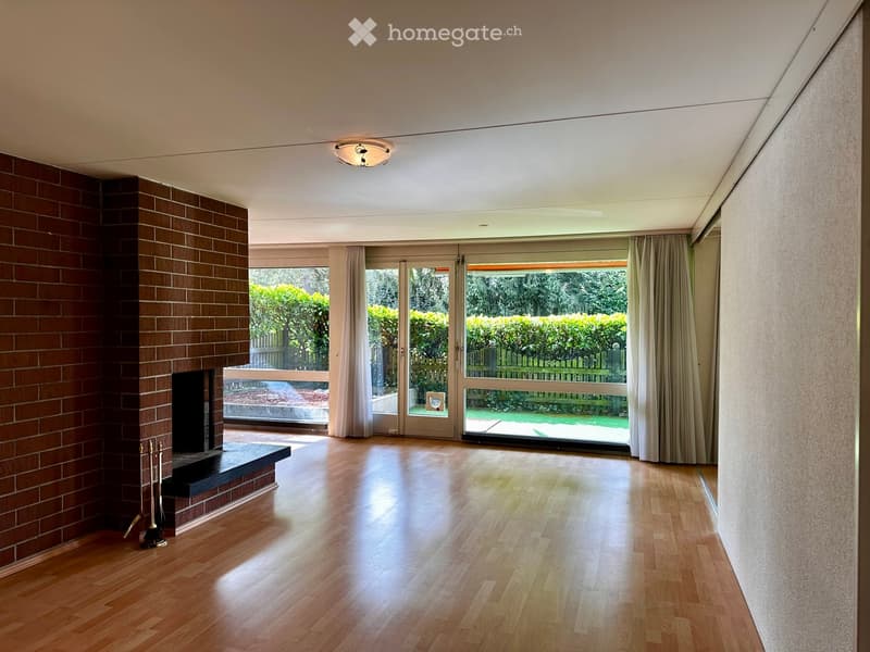Bright living room with wooden flooring, large windows, and a brick fireplace. The space is open and well-lit, with a view of a garden through the windows.