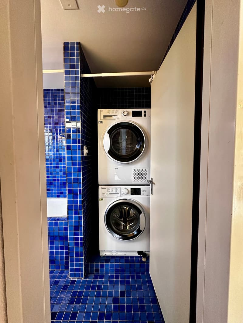 A small bathroom with blue tiled walls and floor, featuring a white washing machine and dryer unit.