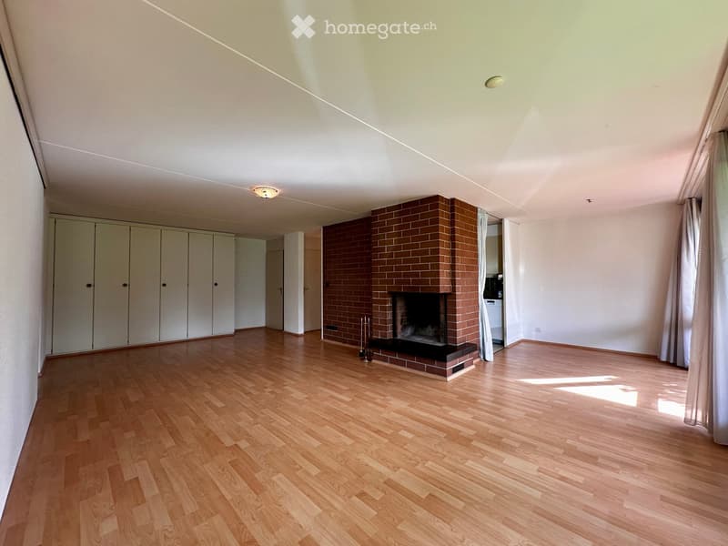Empty room with wooden flooring, white walls, and a brick fireplace. There are built-in white cabinets on one side and a curtained window on the opposite side.