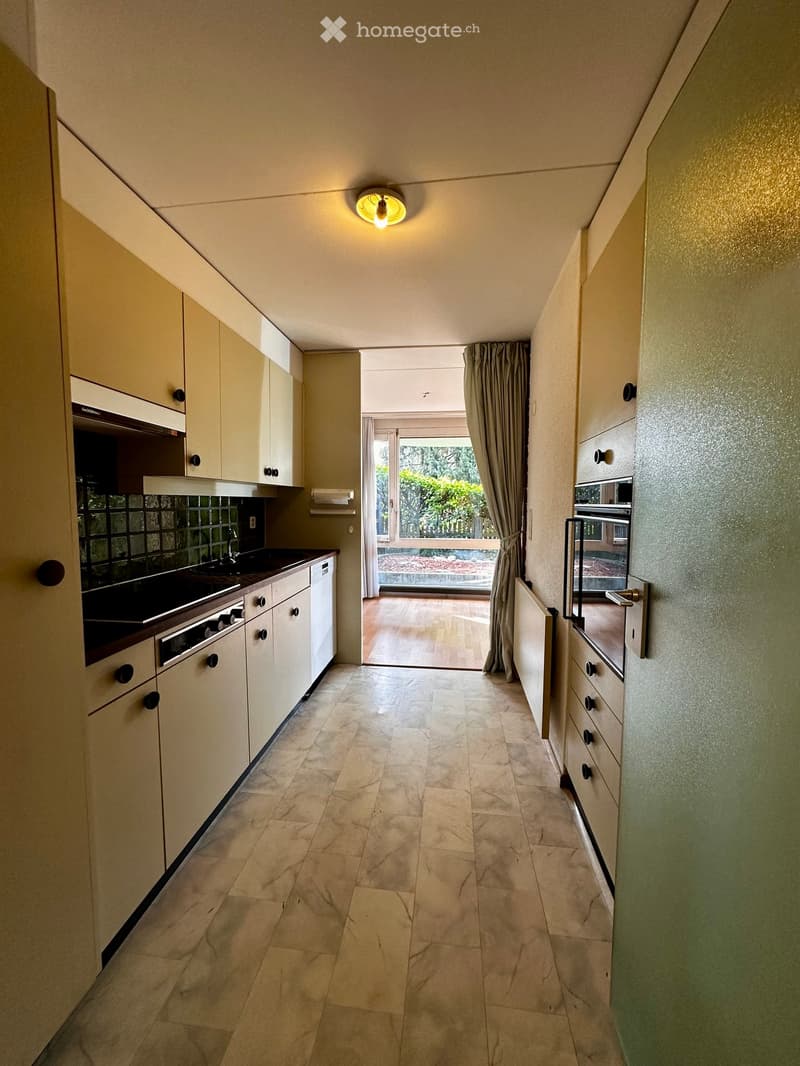 A narrow hallway with light-colored cabinets on both sides, leading to a kitchen area with a sink, stove, and oven. The floor is tiled, and there is a curtained doorway at the end.