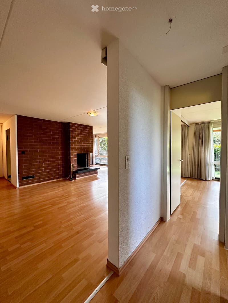 A well-lit hallway with wooden flooring, white walls, and a visible fireplace in an adjacent room. The hallway features a light fixture on the ceiling and a door leading to another room with curtains.