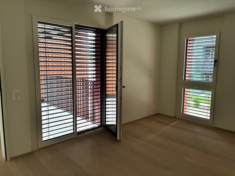 Empty room with light-colored walls, wooden flooring, and large windows with blinds. The room has a sliding glass door leading to a balcony or outdoor area.