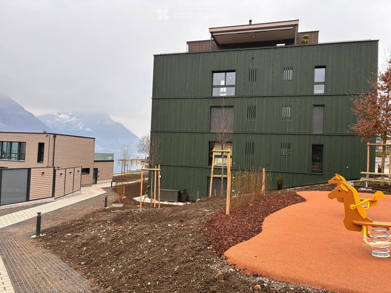 Modern two-story building with green siding and large windows, surrounded by landscaped area with playground equipment and a scenic mountain view.