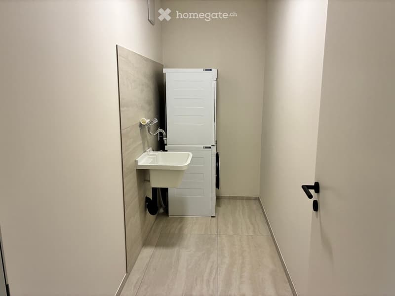 Small bathroom with a white sink, faucet, toilet paper holder, and a white washing machine. Light-colored tiled walls and floor.
