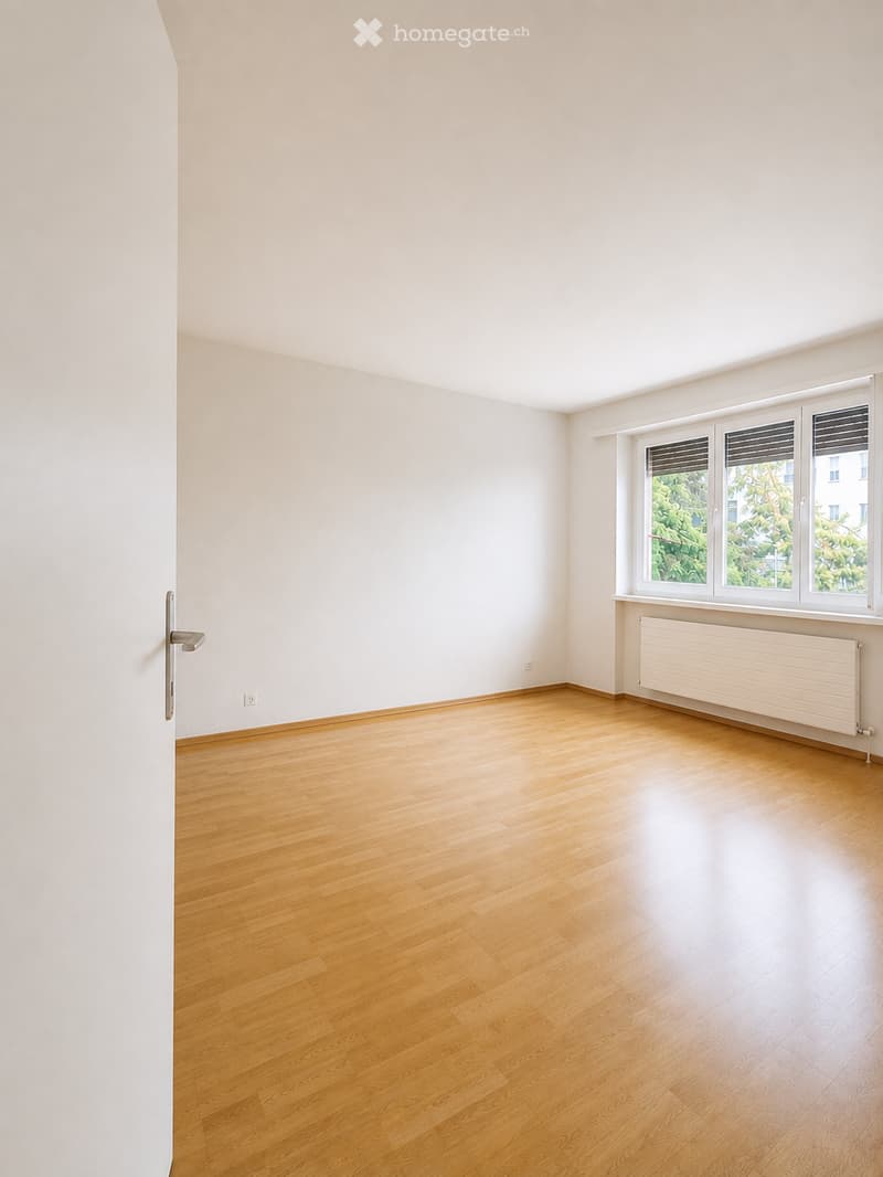 Empty room with light wooden flooring, white walls, and large windows with blinds. No furniture or equipment present.