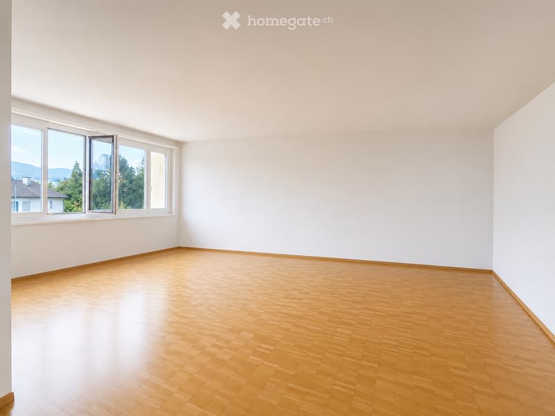 Empty room with light wooden flooring and white walls, featuring large windows with a scenic view of trees and mountains.