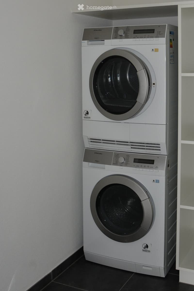 Stacked washer and dryer appliances, dishwasher below, white appliances against a white wall, black tiled floor